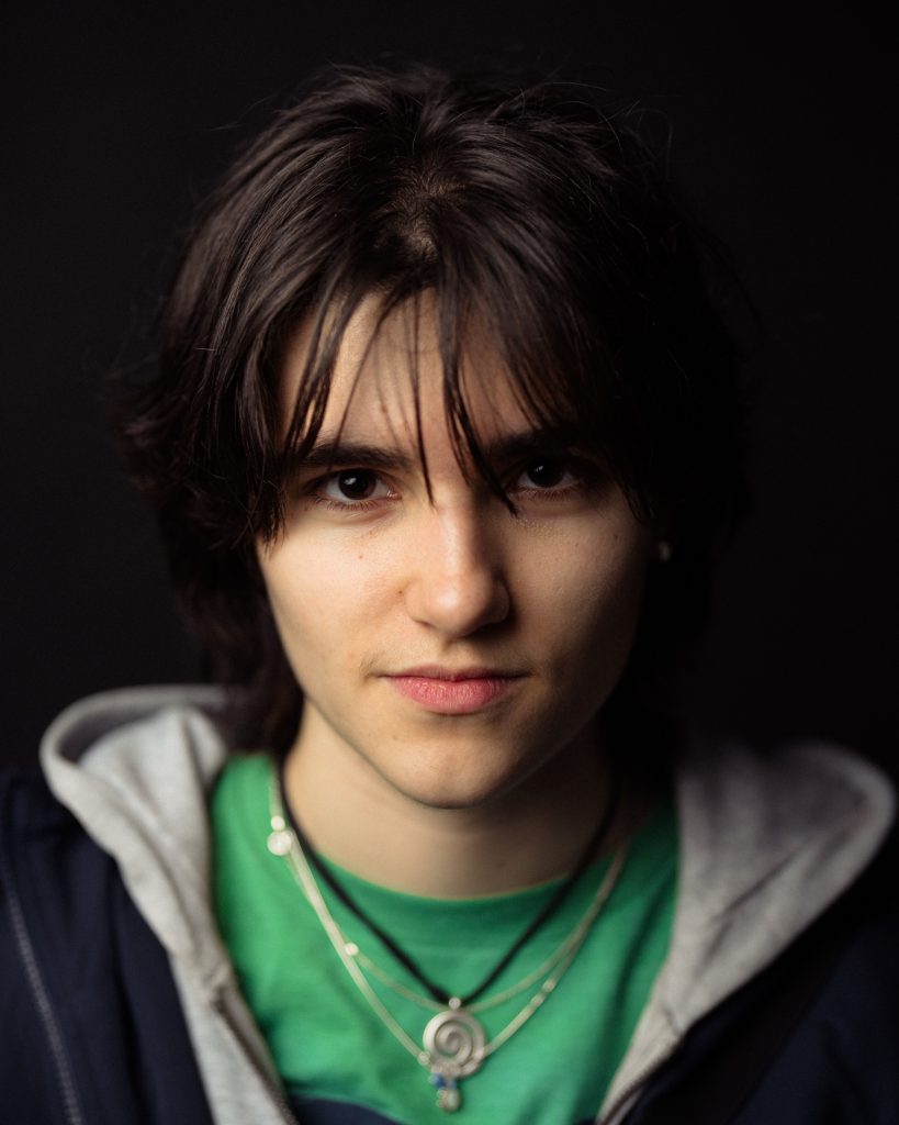 Portrait of a young woman with short dark hair, wearing a green shirt and layered necklaces, against.