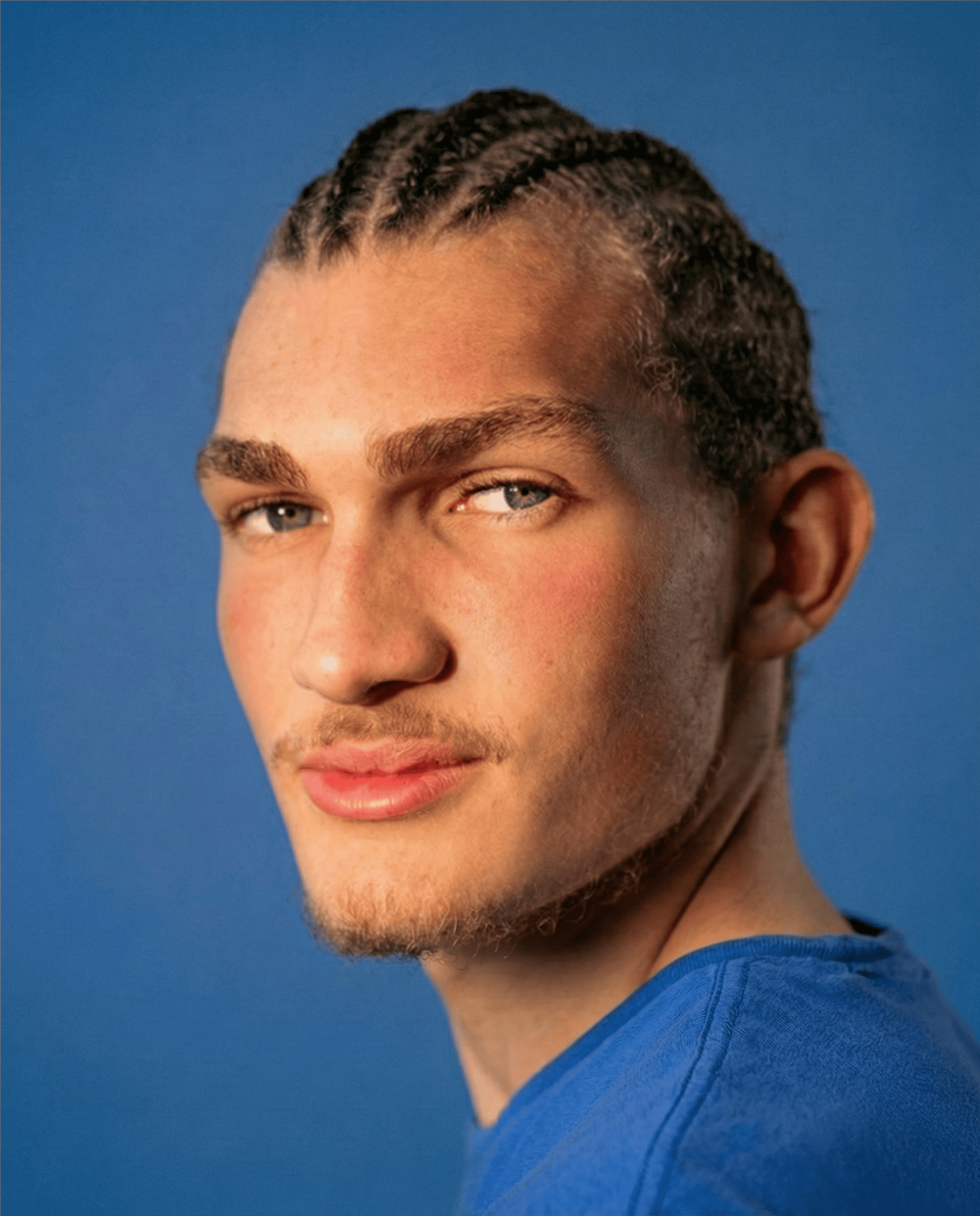 Studio portrait of a young man with cornrow braids wearing a blue t-shirt against a cobalt blue background, LemonLens portrait photography London
